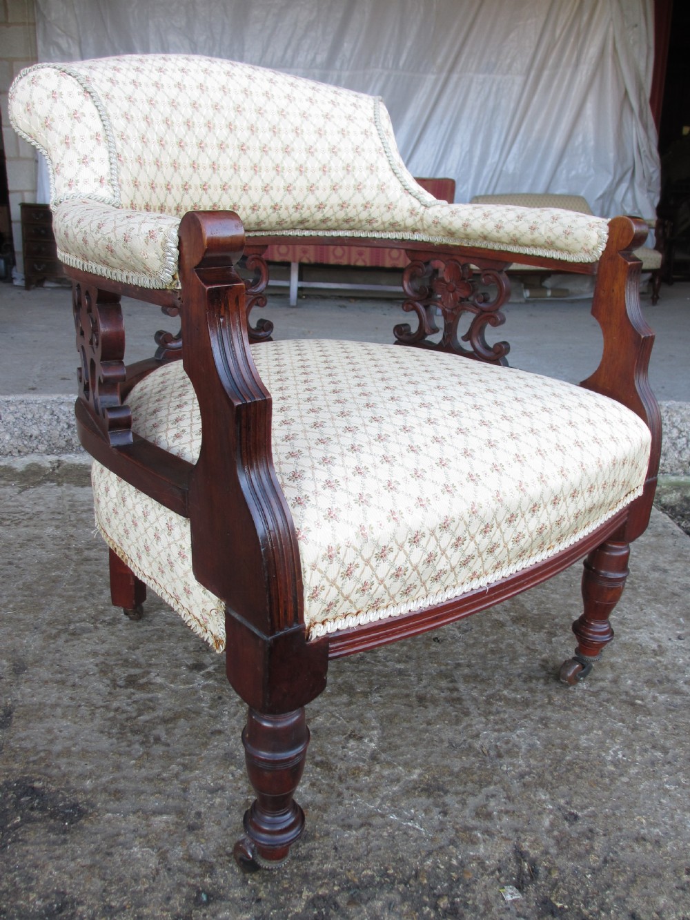 A pair of antique mahogany tub armchairs featuring cream patterned fabric upholstery, openwork carved back splats, and turned front legs on castors. View V
