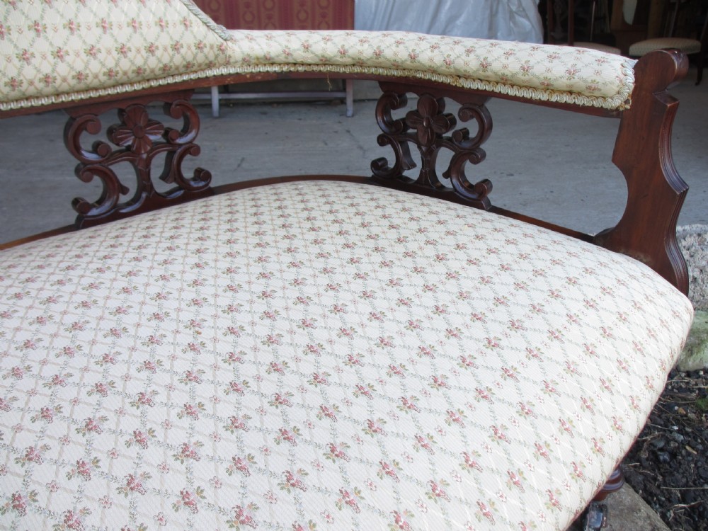 An Edwardian mahogany settee featuring a padded backrest and seat upholstered in cream patterned fabric, supported by carved back splats and turned front legs on castors. View V
