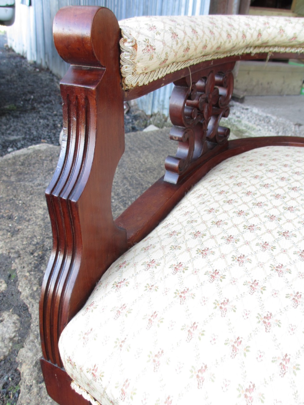 An Edwardian mahogany settee featuring a padded backrest and seat upholstered in cream patterned fabric, supported by carved back splats and turned front legs on castors. View VI