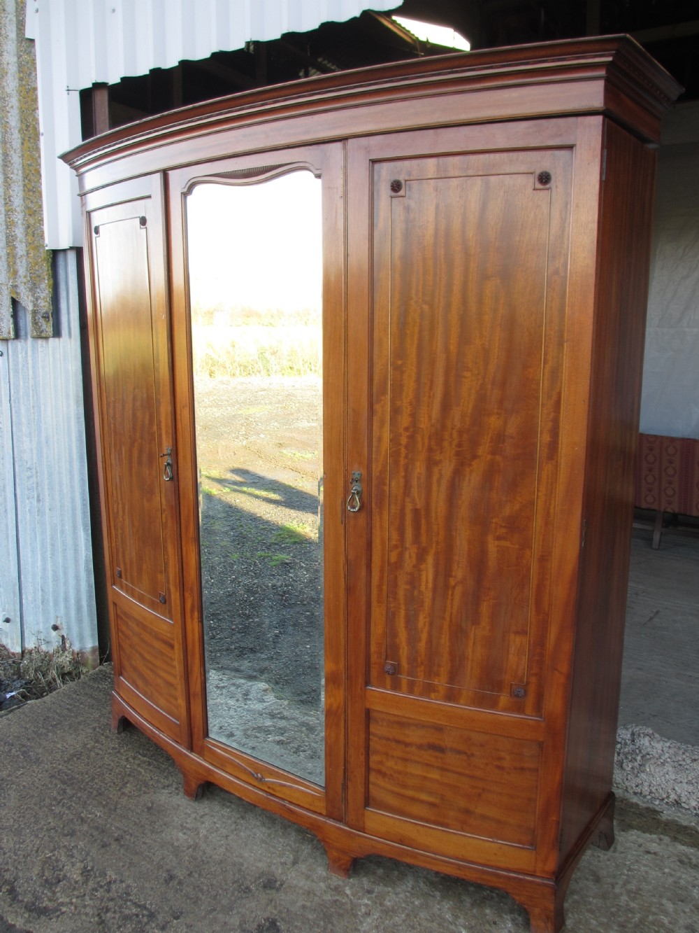 A Victorian bow-fronted mahogany triple wardrobe featuring a central full-length mirror door flanked by panelled flame mahogany doors. View II