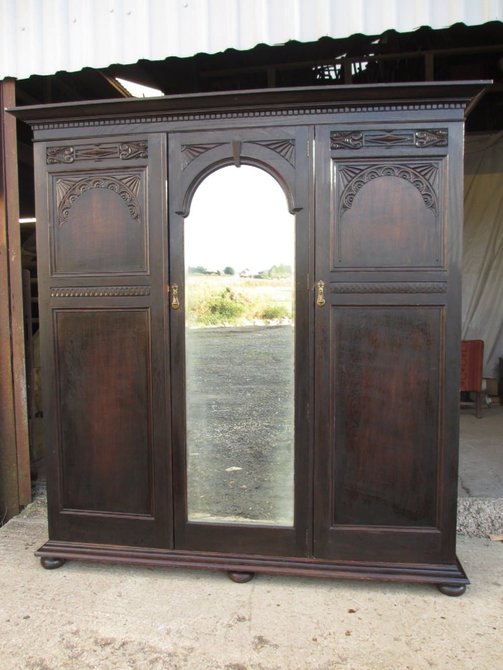 An Edwardian dark oak triple wardrobe featuring carved decorative panels on the side doors, an arched mirrored centre door, and flattened bun feet.