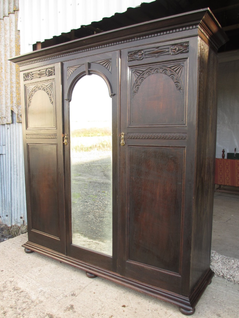 An Edwardian dark oak triple wardrobe featuring carved decorative panels on the side doors, an arched mirrored centre door, and flattened bun feet. View II