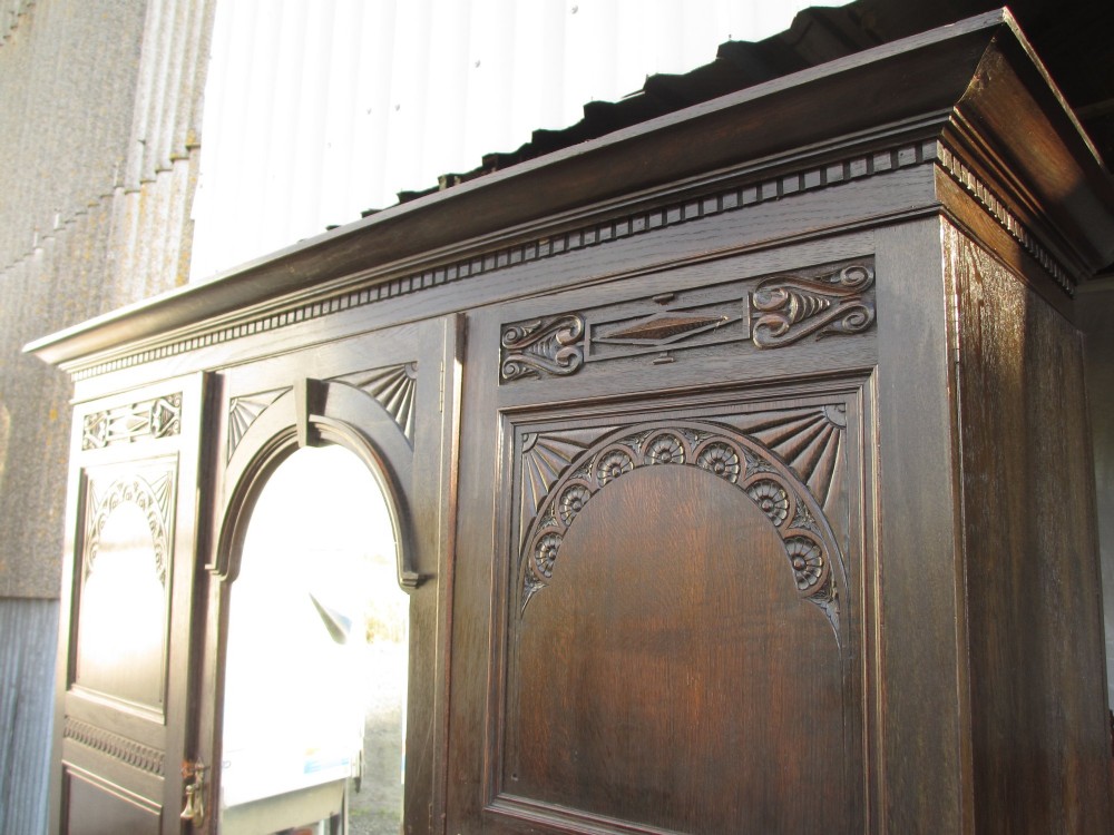 An Edwardian dark oak triple wardrobe featuring carved decorative panels on the side doors, an arched mirrored centre door, and flattened bun feet. View III