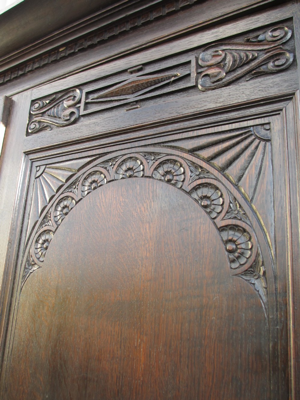 An Edwardian dark oak triple wardrobe featuring carved decorative panels on the side doors, an arched mirrored centre door, and flattened bun feet. View V