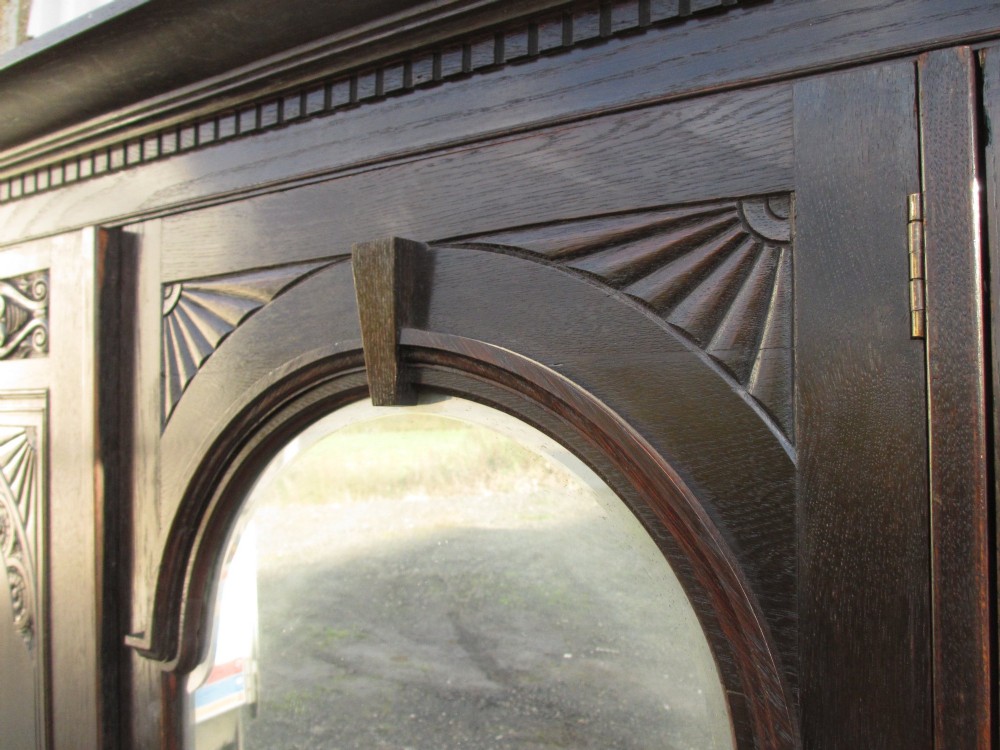 An Edwardian dark oak triple wardrobe featuring carved decorative panels on the side doors, an arched mirrored centre door, and flattened bun feet. View VII