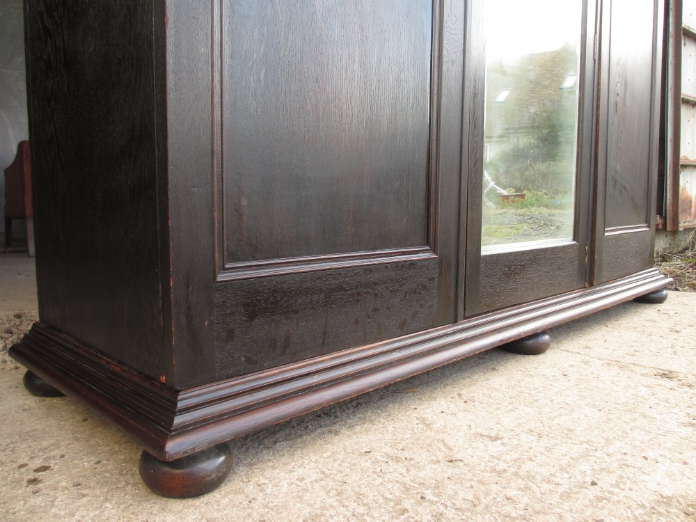 An Edwardian dark oak triple wardrobe featuring carved decorative panels on the side doors, an arched mirrored centre door, and flattened bun feet. View VIII