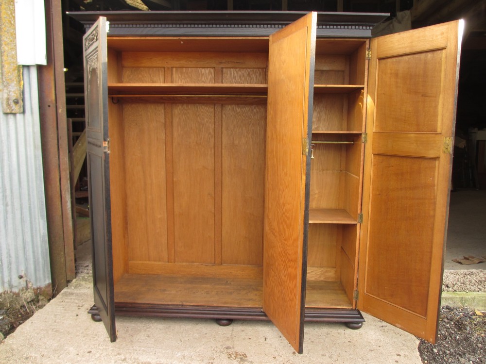 An Edwardian dark oak triple wardrobe featuring carved decorative panels on the side doors, an arched mirrored centre door, and flattened bun feet. View X