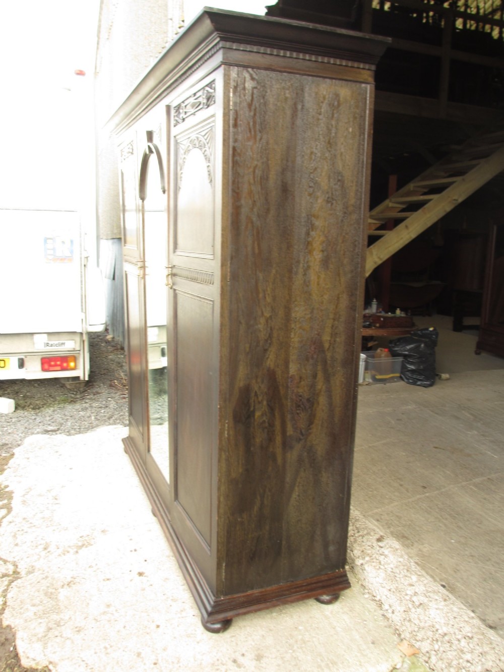 An Edwardian dark oak triple wardrobe featuring carved decorative panels on the side doors, an arched mirrored centre door, and flattened bun feet. View XIV