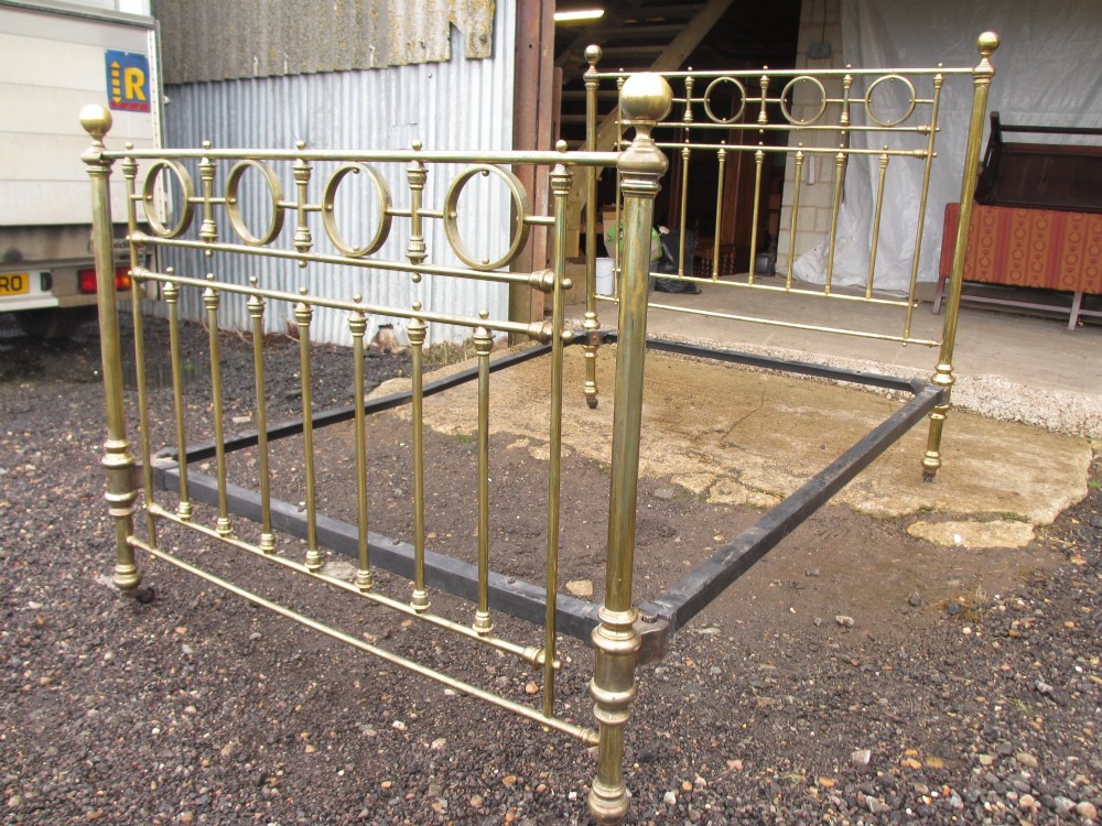 A Victorian brass bed frame featuring circular motifs, vertical spindles, ball finials, and an iron slot frame set on castors. View II