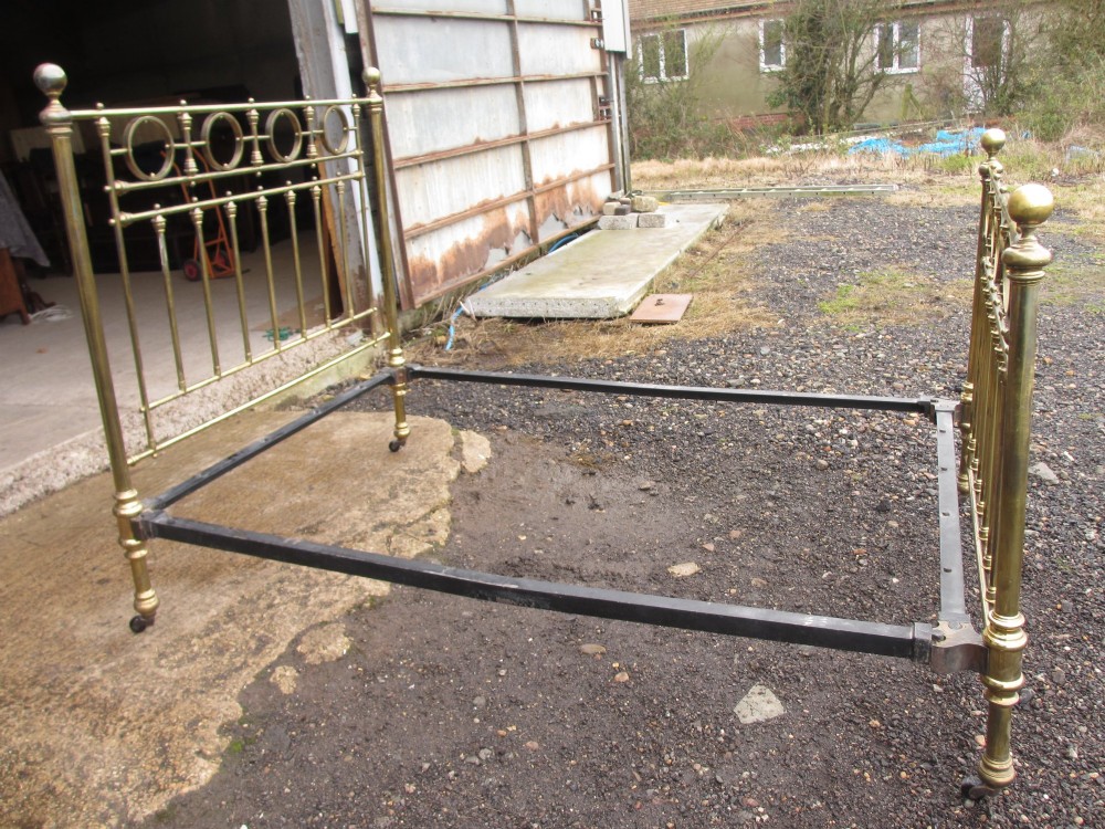 A Victorian brass bed frame featuring circular motifs, vertical spindles, ball finials, and an iron slot frame set on castors. View V