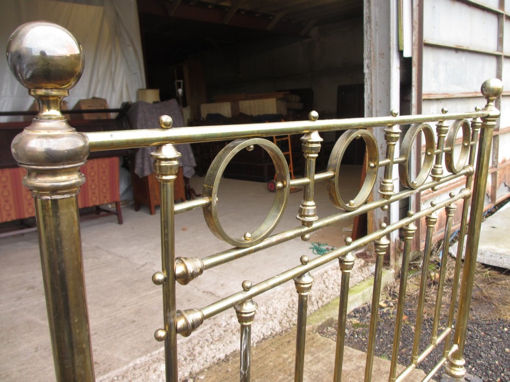 A Victorian brass bed frame featuring circular motifs, vertical spindles, ball finials, and an iron slot frame set on castors. View VI