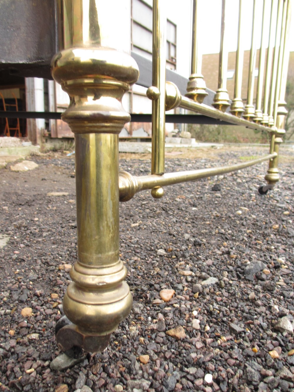 A Victorian brass bed frame featuring circular motifs, vertical spindles, ball finials, and an iron slot frame set on castors. View X