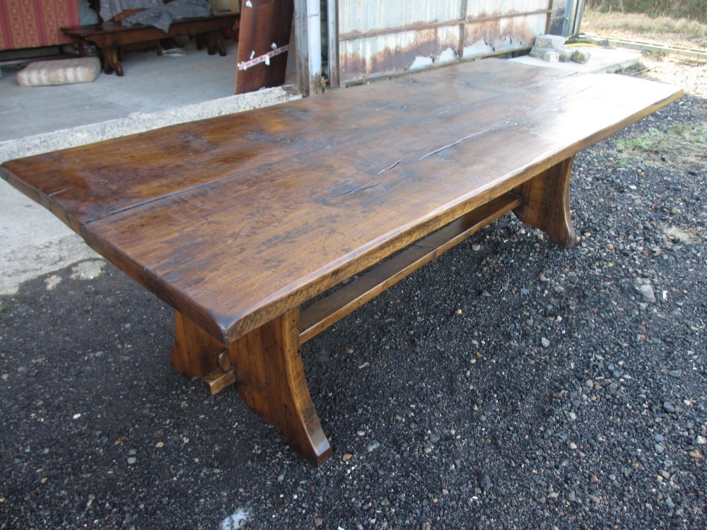 A solid oak refectory dining table featuring a thick plank top, shaped trestle ends, and a low central stretcher. View IV