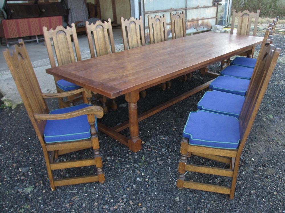 A long twentieth-century oak refectory dining table with turned legs and stretcher base, accompanied by twelve high-backed carved oak dining chairs with blue seat cushions. View XIII