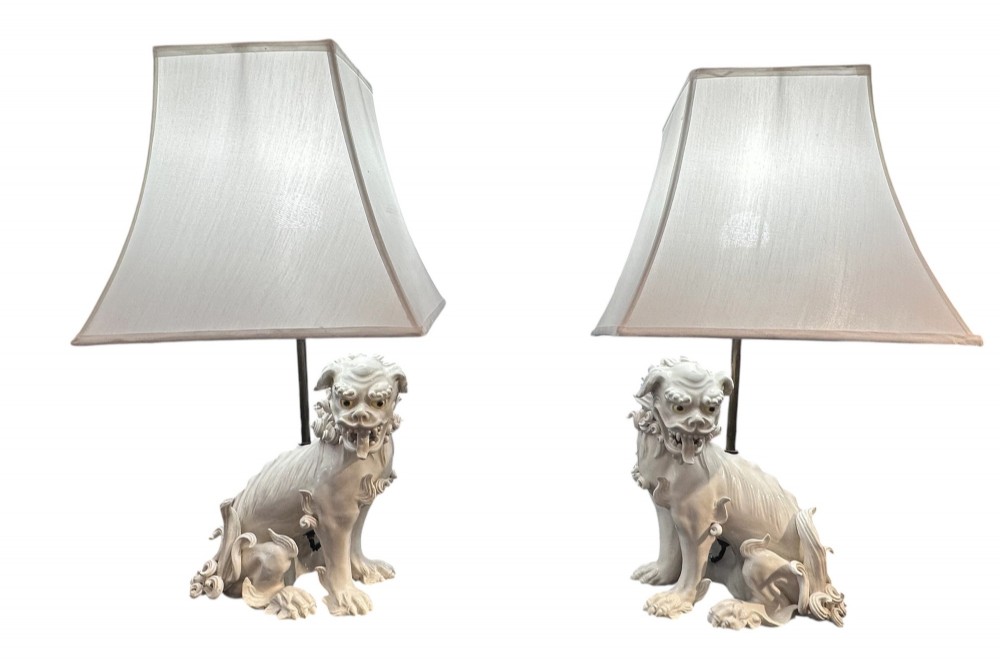 A pair of early 20th-century off-white porcelain Foo Dog table lamps with tall cream fabric shades.